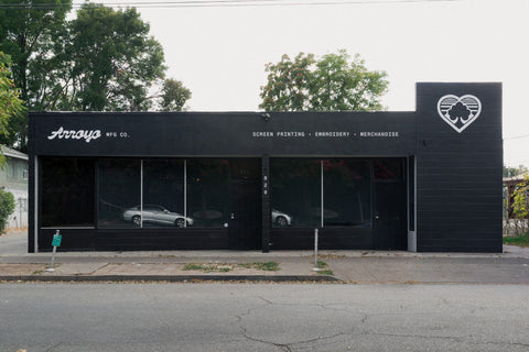 A storefront building with a black facade and large glass windows, featuring the brand name 'Arrays' and the text 'SCREEN PRINTING • EMBROIDERY • MERCHANDISE.' Trees are visible above the building, and a street and sidewalk are in front. Arroyo MFG Co. custom apparel shop exterior - 820 Wall St Chico Ca