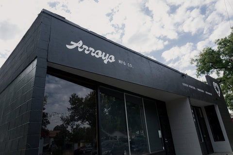 The exterior of the Arrows MFG Co. building, featuring a dark gray facade, large glass windows, and white script branding on the upper wall under a cloudy sky. Arroyo MFG Co. custom apparel shop exterior - 820 Wall St Chico Ca