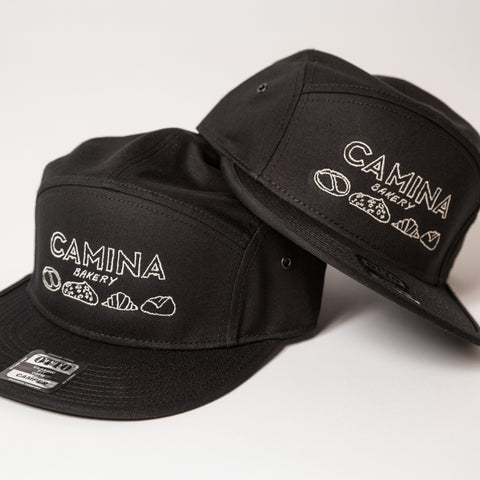 Two black caps with embroidery 'Camina Bakery' branding on a white background