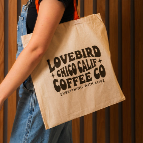 Person holding a beige tote bag with 'Lovebird Chico Calif Coffee Co' text against a wooden background